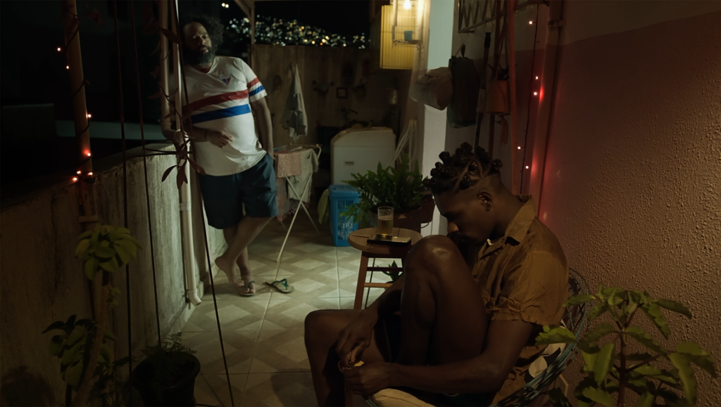 Scene from The Day I Met You film. Two men on a balcony at night, one seated, one standing.