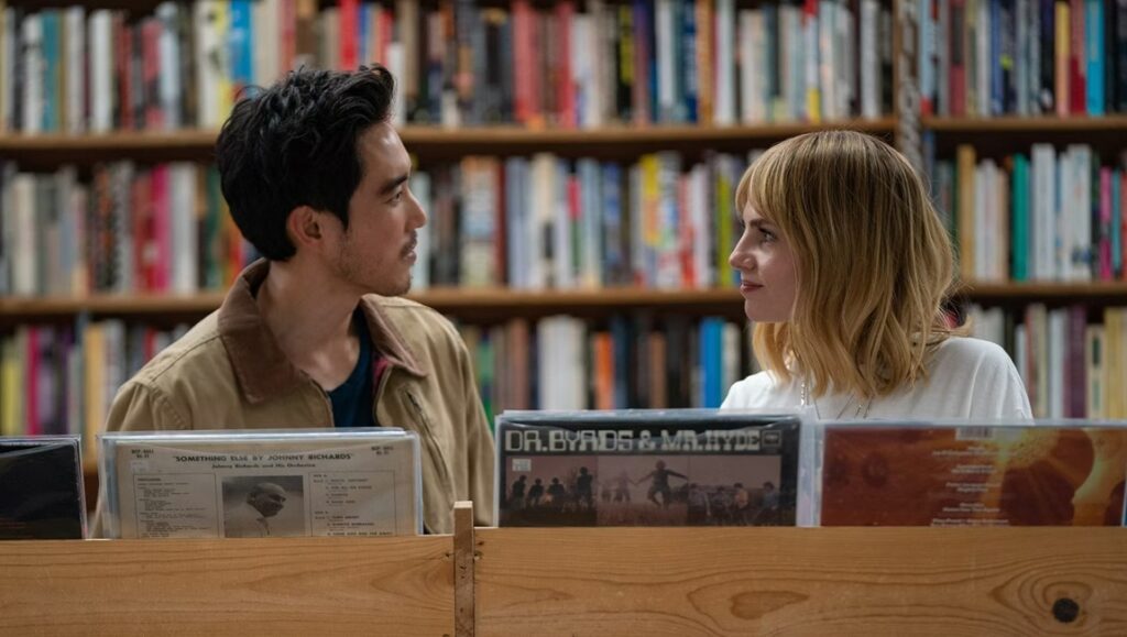 Lucy Boynton and Justin H. Min in The Greatest Hits movie. Couple looking at each other over vinyl records.