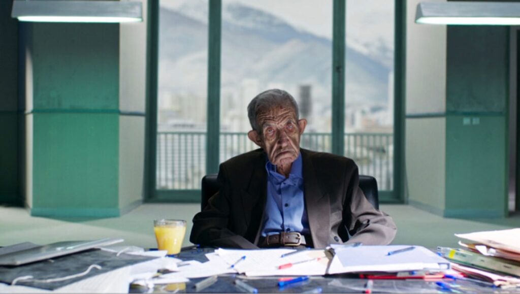 Still from Terrestrial Verses. An older man sits at a desk with papers, pens, and a glass of juice. Mountains in the background.