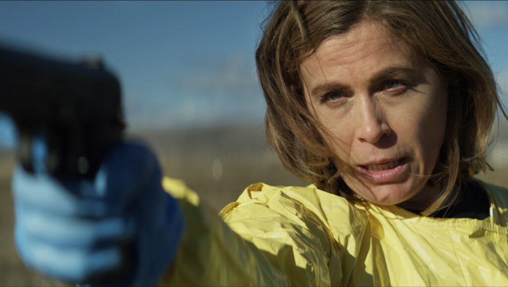 Chelsea Gilligan in New Life movie review. Woman in hazmat suit aims a gun.