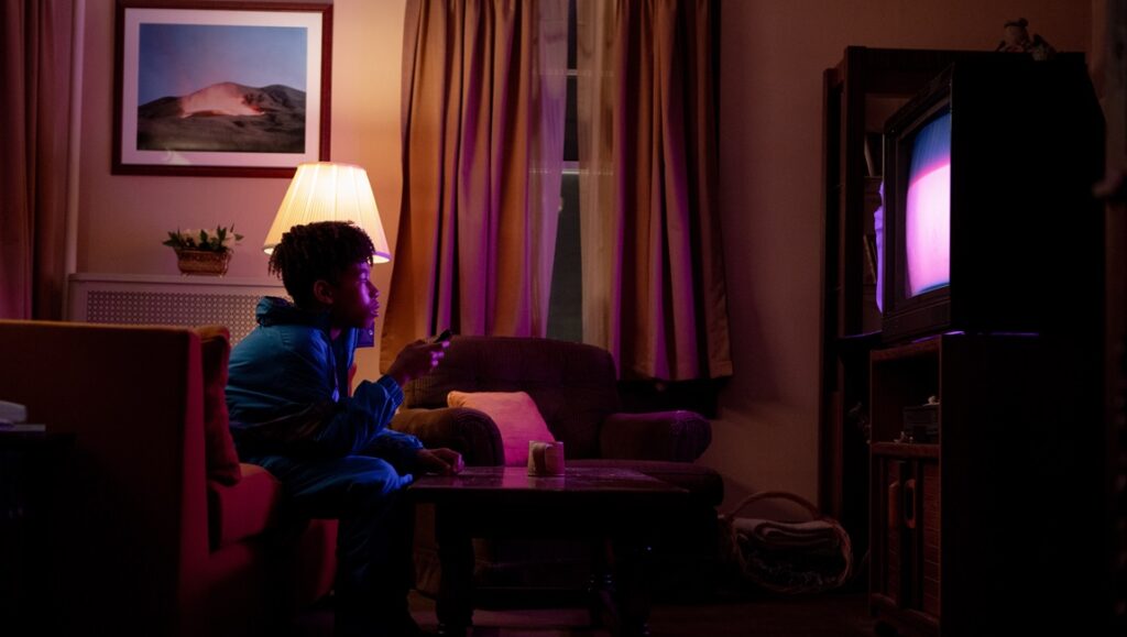 I Saw the TV Glow movie scene: Teen watches TV in a dimly lit room, bathed in the screen's glow. Curtains and a lamp add to the ambiance.