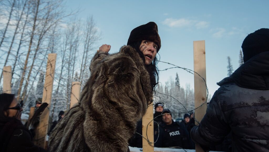 Scene from 2024 Hot Docs Film Festival. Indigenous woman in fur coat confronts police. Barbed wire fence. Protest.