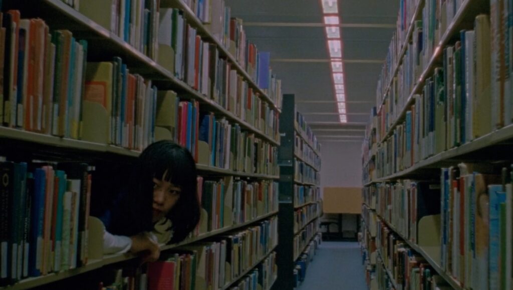 Us and the Night film still: Audrey Lam peers from library bookshelves in Prismatic Ground '24 review.
