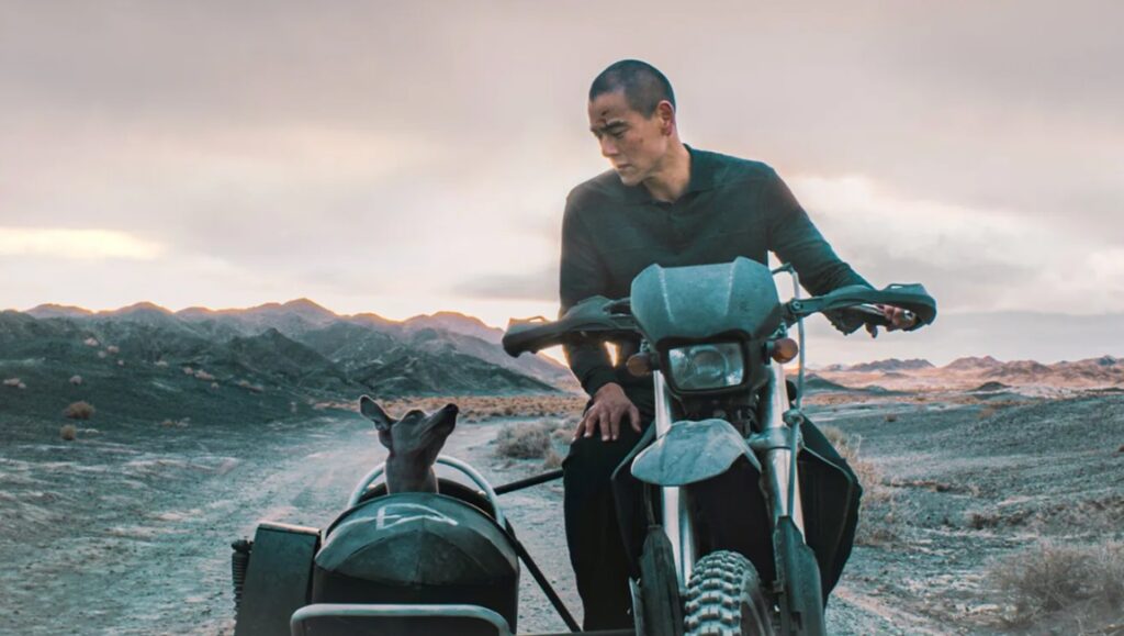 Black Dog film still from Cannes 2024. Man on motorcycle with dog in sidecar, desert landscape.
