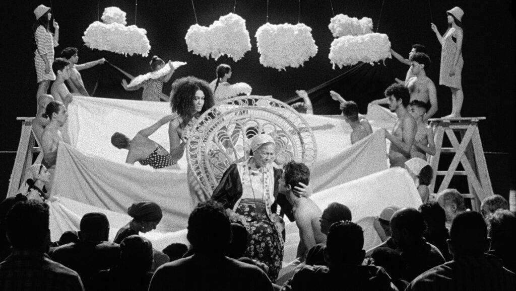 Black and white performance art. Audience watches actors, clouds, and props on stage.