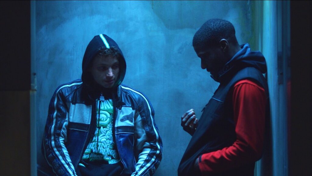 Eat The Night film still, Cannes Film Festival 2024. Two men in hoodies in a blue-lit scene.