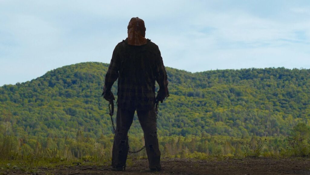 In a Violent Nature movie still. Back view of a masked killer holding chains in front of a wooded mountain landscape.