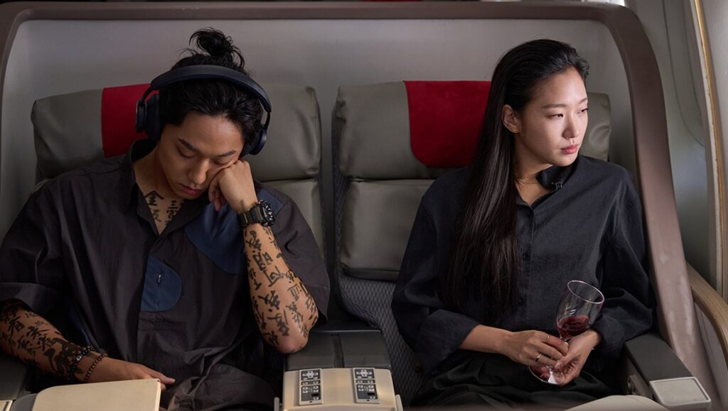 Exhuma Korean horror film scene. Tattooed shaman asleep on a plane, next to a woman with wine.
