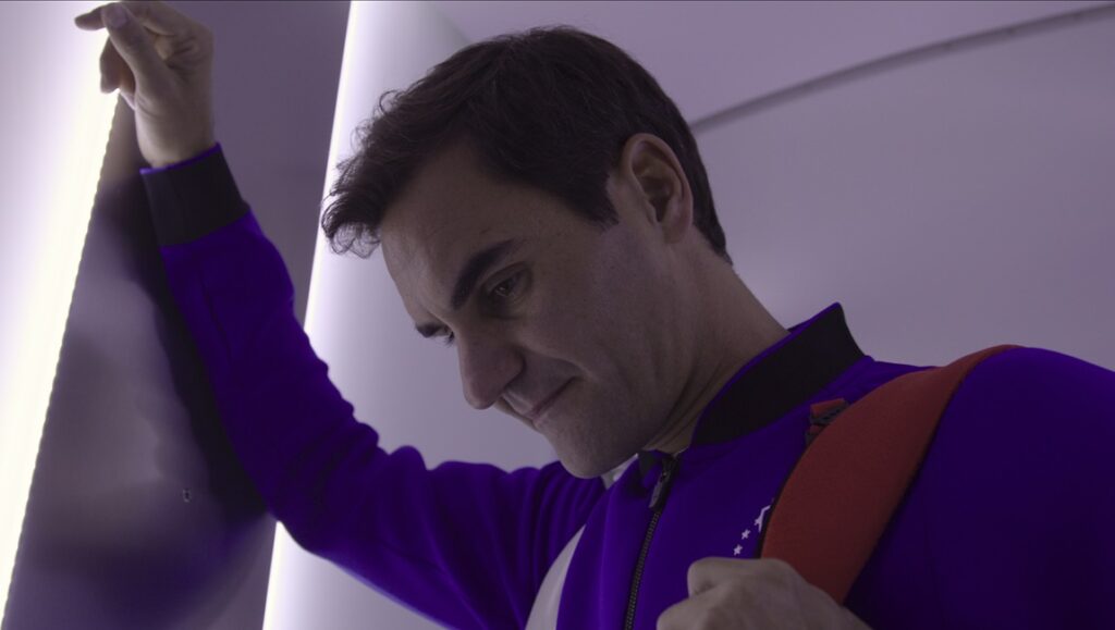 Roger Federer in purple jacket with orange strap, leaning on a wall. Tennis star.