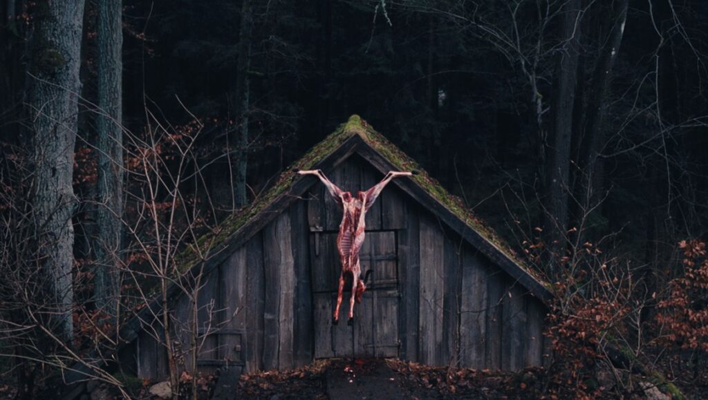 The Devil's Bath movie still: Carcass hangs on a rustic wooden shed in a dark forest. Horror film imagery.