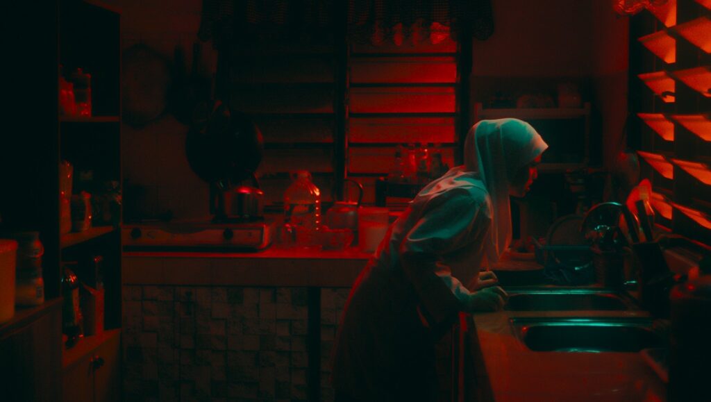 Photogram from Tiger Stripes film. Woman in hijab in a dimly lit kitchen with red lighting. Supernatural horror.