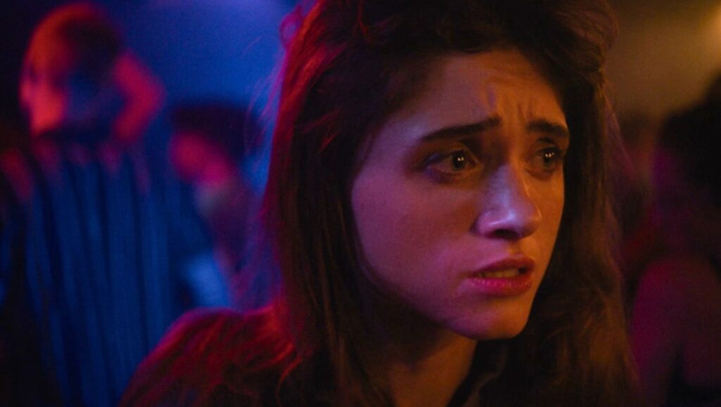 Chestnut movie scene: Natalia Dyer with a concerned expression in a crowded, dimly lit setting.