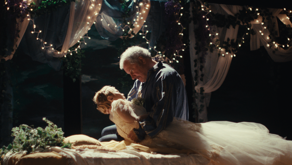 Ghostlight movie scene: older man embraces woman in white dress on a stage set with lights and greenery.