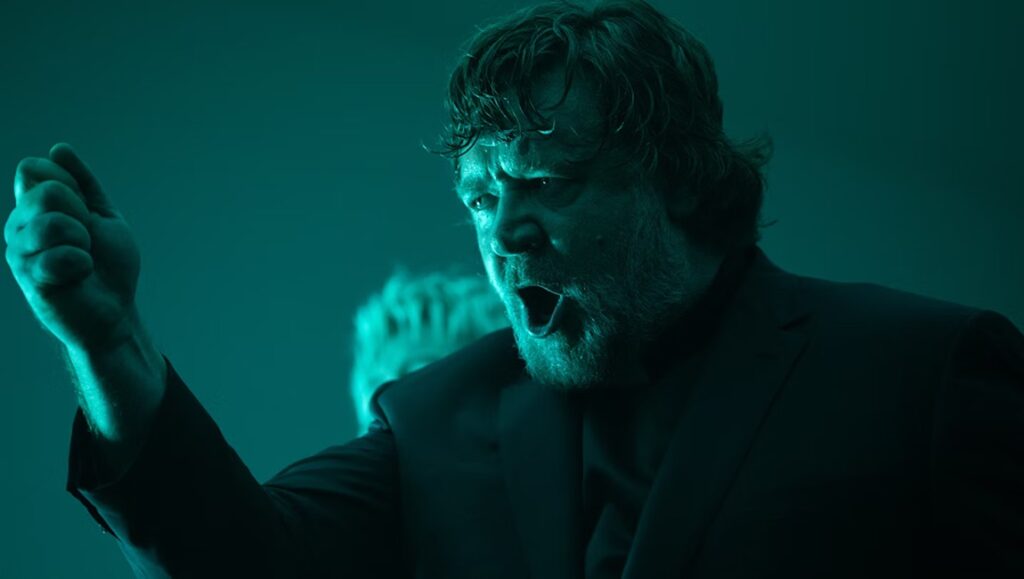 Russell Crowe in The Exorcism movie. Actor with beard, mouth open, and fist raised. Horror film review.