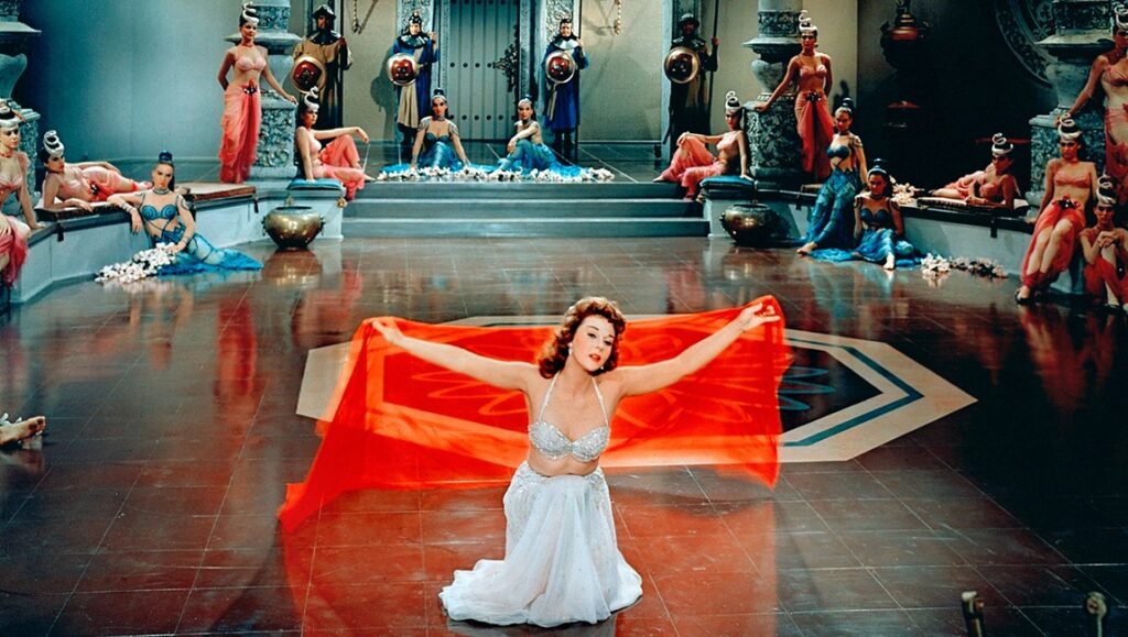 Still from The Conqueror movie. Woman dancing with red fabric, surrounded by women and guards.