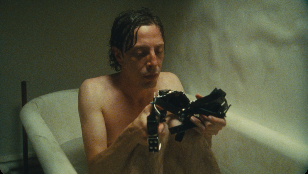 Naked man in bathtub holds a strange device. Dark hair, pale skin. Bizarre.