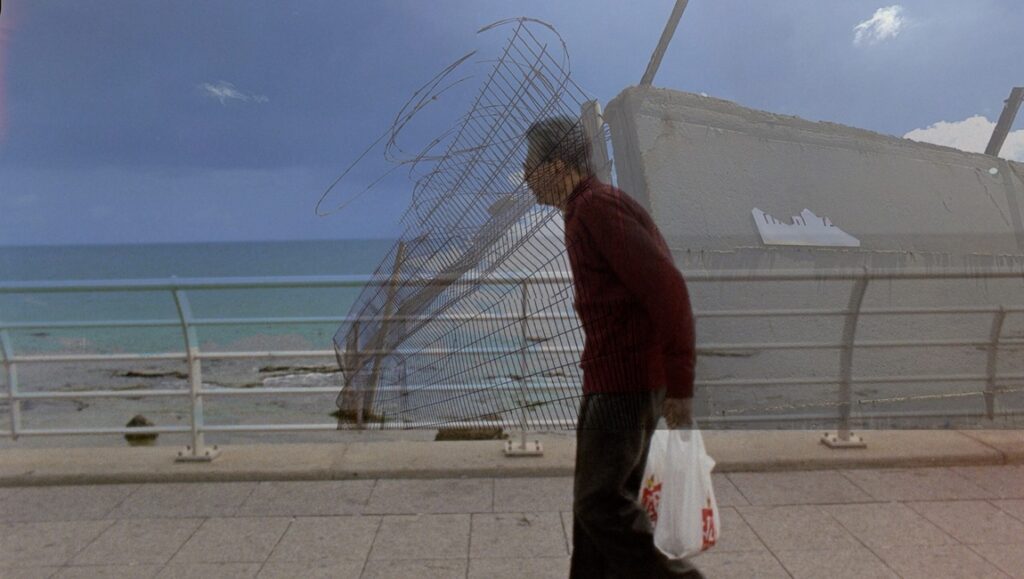 Film still from Jessica Dunn Rovinelli's Life Story (FIDMarseille '24). Man walks by the sea, carrying a bag.