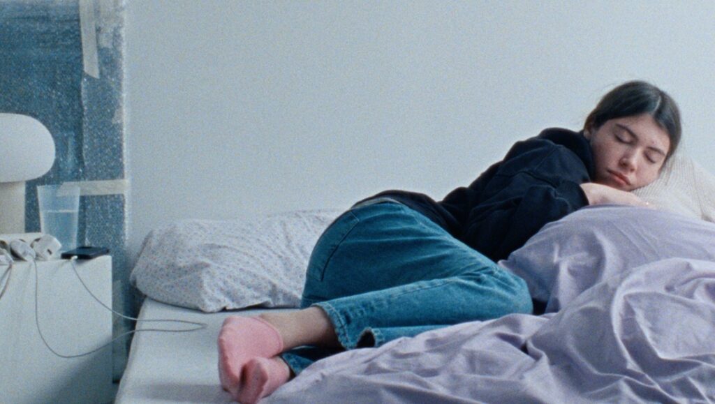 Still from Lazaro at Night FIDMarseille 2024 film. Young woman sleeping in bed, wearing jeans and a hoodie. Indie film scene.
