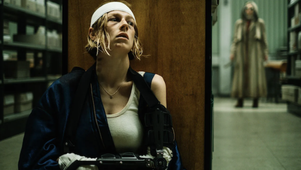 Hunter Schafer in Cuckoo horror movie. Woman with head bandage, holding a gun, and a figure in the background.