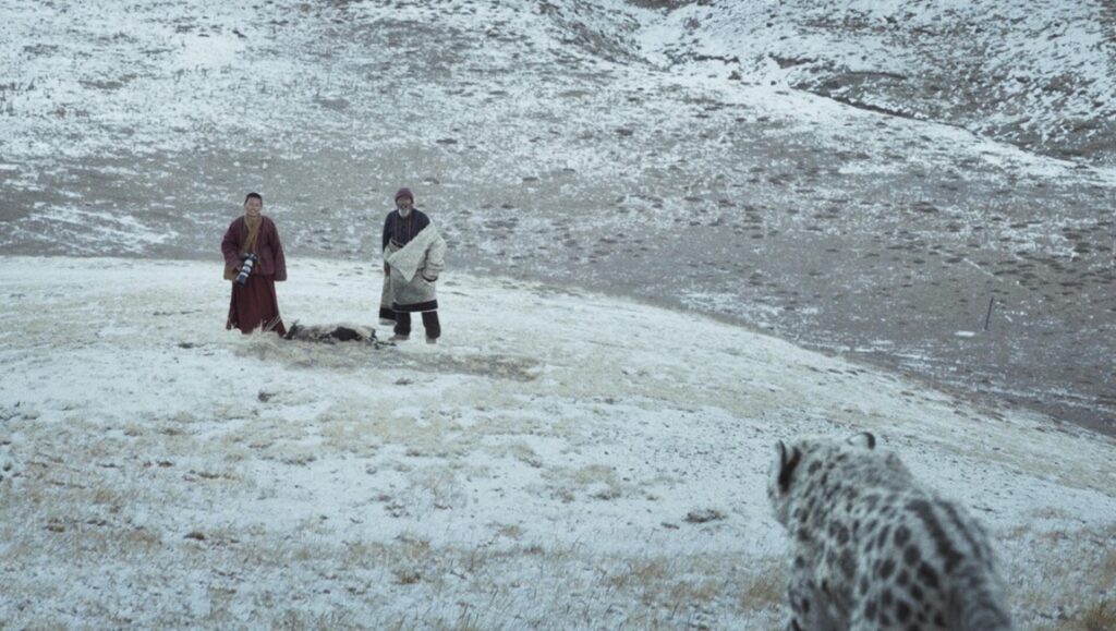 Snow Leopard NYAFF film still. Two figures stand near a carcass as a snow leopard approaches.