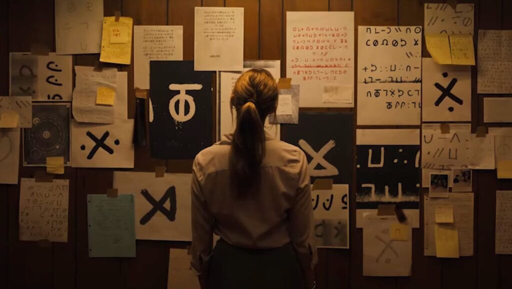 Longlegs movie scene: Woman facing a wall covered in symbols and cryptic notes. Horror film imagery.