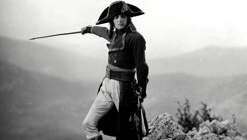 Black and white still from Abel Gance's Napoleon film. Napoleon stands with a sword on a rocky outcrop.