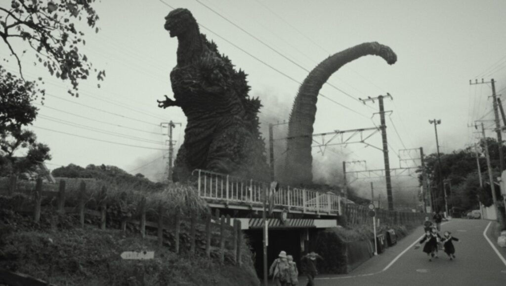 Black and white still from Shin Godzilla film. Godzilla towers over a bridge as people flee.