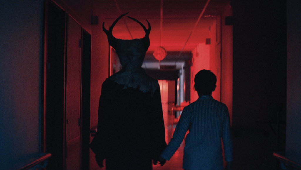 The Soul Eater film still from Fantasia Fest: Horned figure and child walk down a red-lit corridor. Horror.