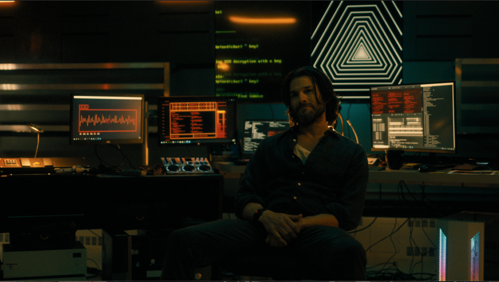 A-Frame film still from Fantasia Fest 2024. Bearded man sits surrounded by computer screens displaying code and data.