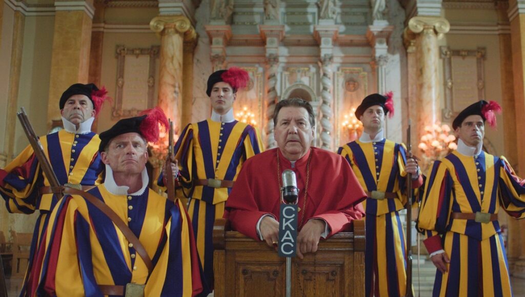 Comedy scene: Cardinal speaks at a mic, flanked by Swiss Guards in traditional yellow and blue striped uniforms.