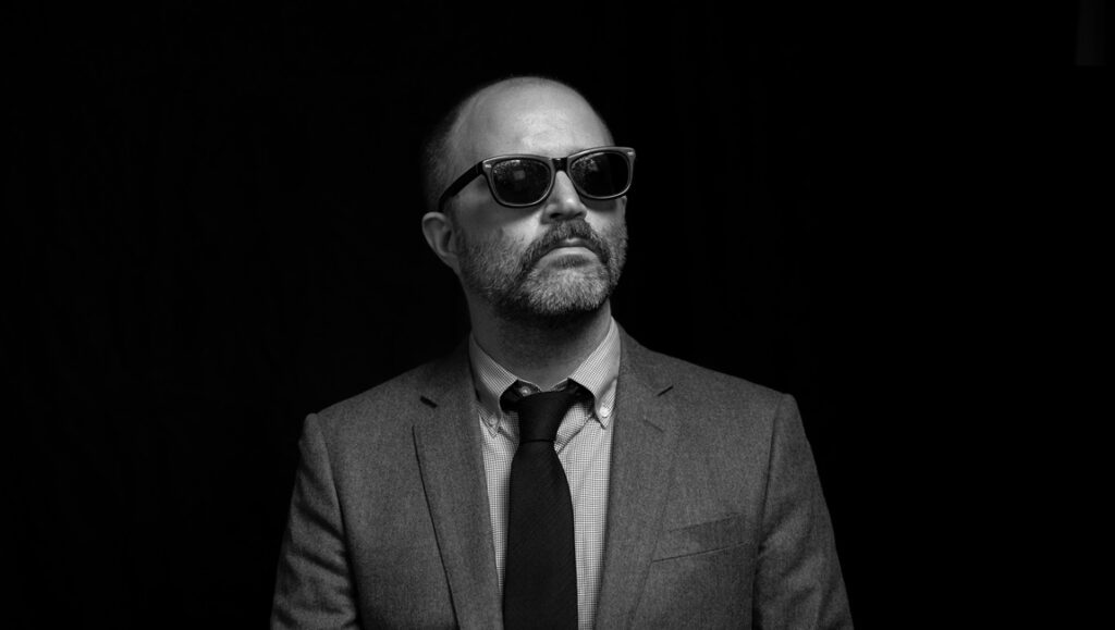 Zach Clark portrait. Black and white photo of Clark in sunglasses, suit, and tie. Interview with Zach Clark.