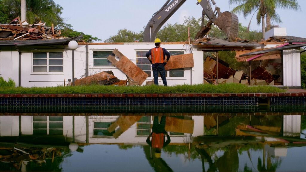 Mountains movie scene: Building demolition with excavator and worker reflected in pool.