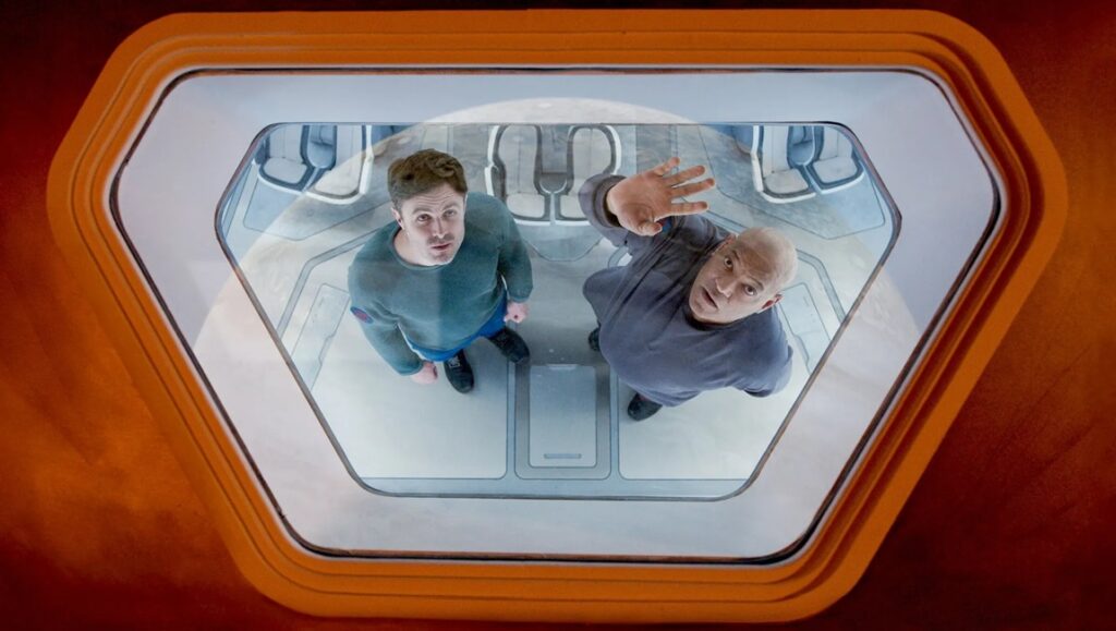 Movie scene from Slingshot featuring two men in a futuristic setting, looking upwards.