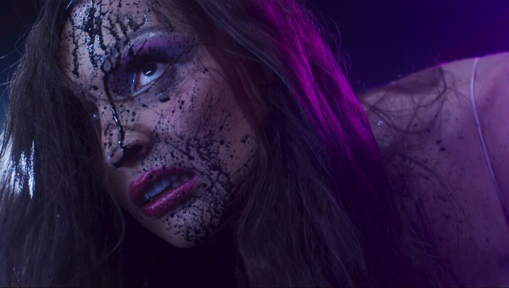 Satranic Panic movie still: Woman with dark makeup, purple eyeshadow, and pink lipstick. Horror film aesthetic.
