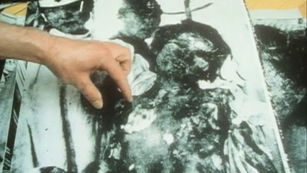 Still from Peter Watkins' film The Journey showing a hand pointing to a black and white photo.