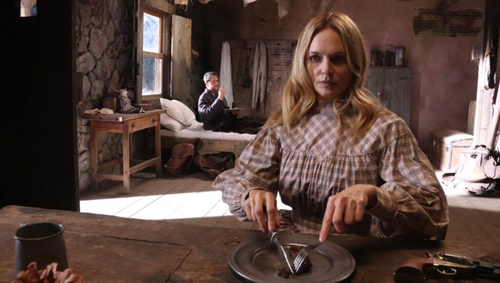 Place of Bones review: Woman eats at table, man sits on bed in rustic cabin. Western horror movie scene.