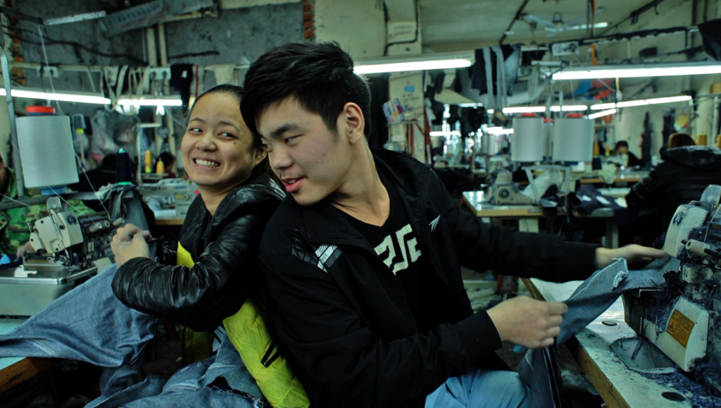 Youth (Hard Times) film still: Two young workers in a Chinese garment factory smile near sewing machines.