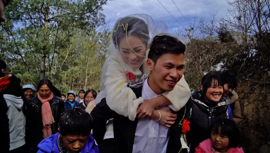 Youth (Homecoming) film still: Groom carries bride piggyback. Wedding celebration, Wang Bing film review.