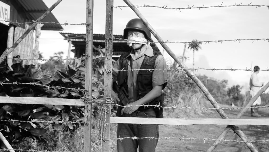 Black and white shot of actor Ronnie Lazaro in Phantosmia, standing behind a barbed wire fence, wearing a helmet.