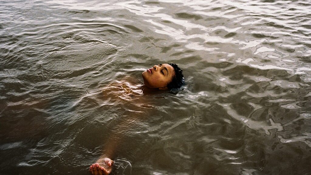 Still from Bird film review: Young person floating in water with eyes closed.