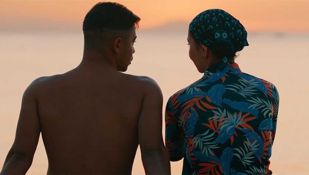 Still from Friendship at TIFF 2024. Two friends at sunset, one shirtless, the other in a floral shirt and headscarf.