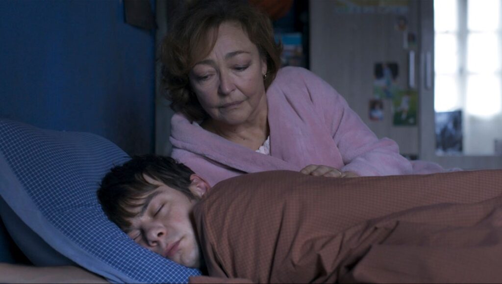 Misericordia film still: A sleeping man is watched over by a concerned woman in a pink robe. Alain Guiraudie film review.