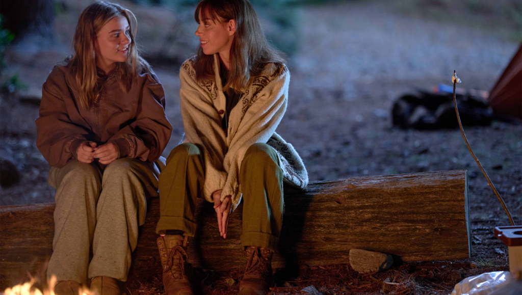 Maisy Stella and Aubrey Plaza in My Old Ass movie. Actresses sit by campfire on a log. Film review.