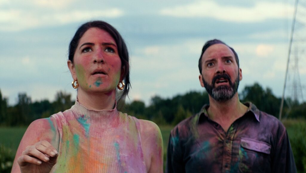 Pepe film review still. Woman and man covered in paint look up with surprise.