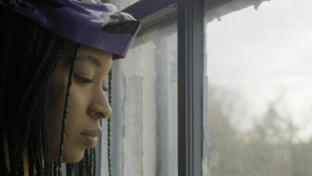 Ghost Game movie still: Woman with braids and mask looks out a rainy window. Jill Gevargizian film review.