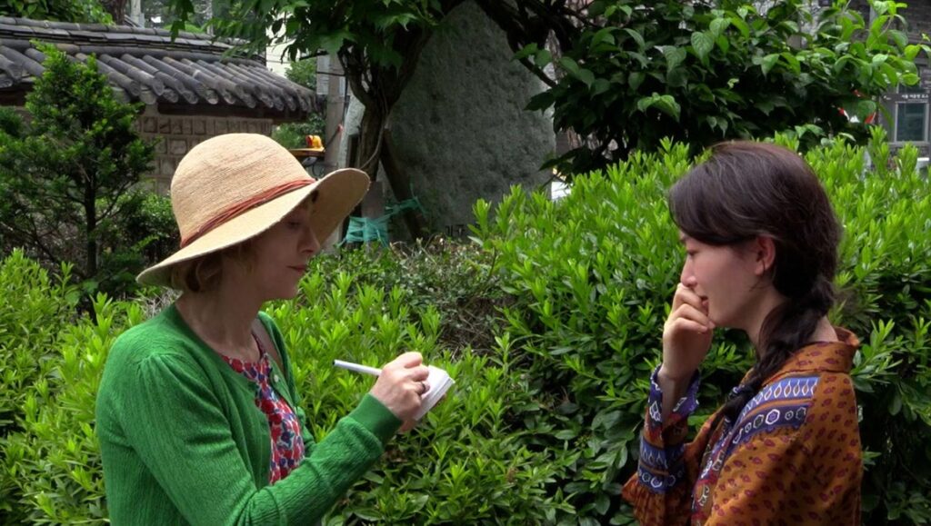 A Traveler's Needs film still. Two women in a garden setting; one takes notes. Hong Sang-soo movie review.