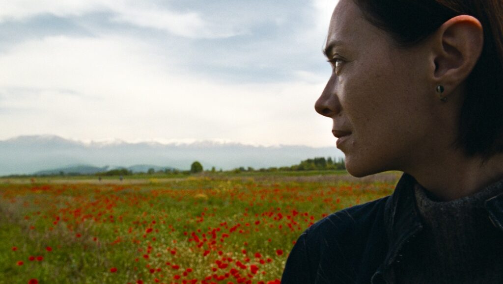 Still from April by Dea Kulumbegashvili. Woman in field of poppies, looking towards distant mountains.