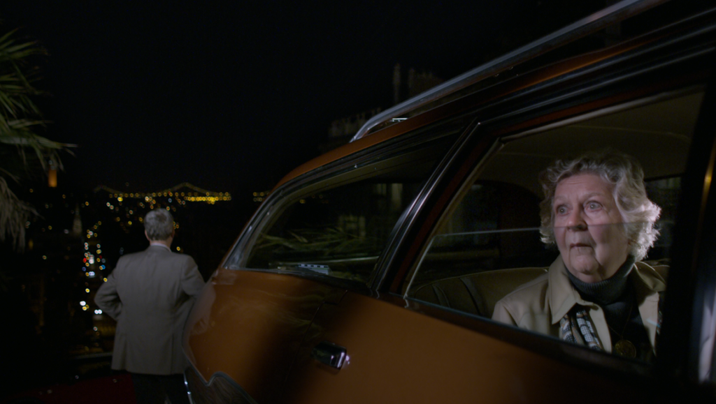 Still from A Traveler's Needs film review. Woman in car, man outside, city lights at night. Hong Sang-soo movie scene.