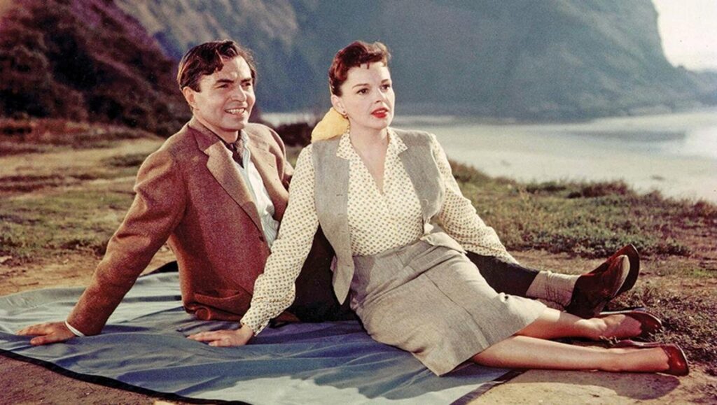 A Star is Born (1954) film still: James Mason and Judy Garland sitting on a blanket outdoors.