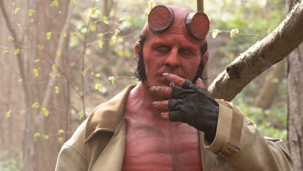 Hellboy in The Crooked Man movie. Red-skinned demon with horns, wearing a coat and fingerless gloves in a forest setting.